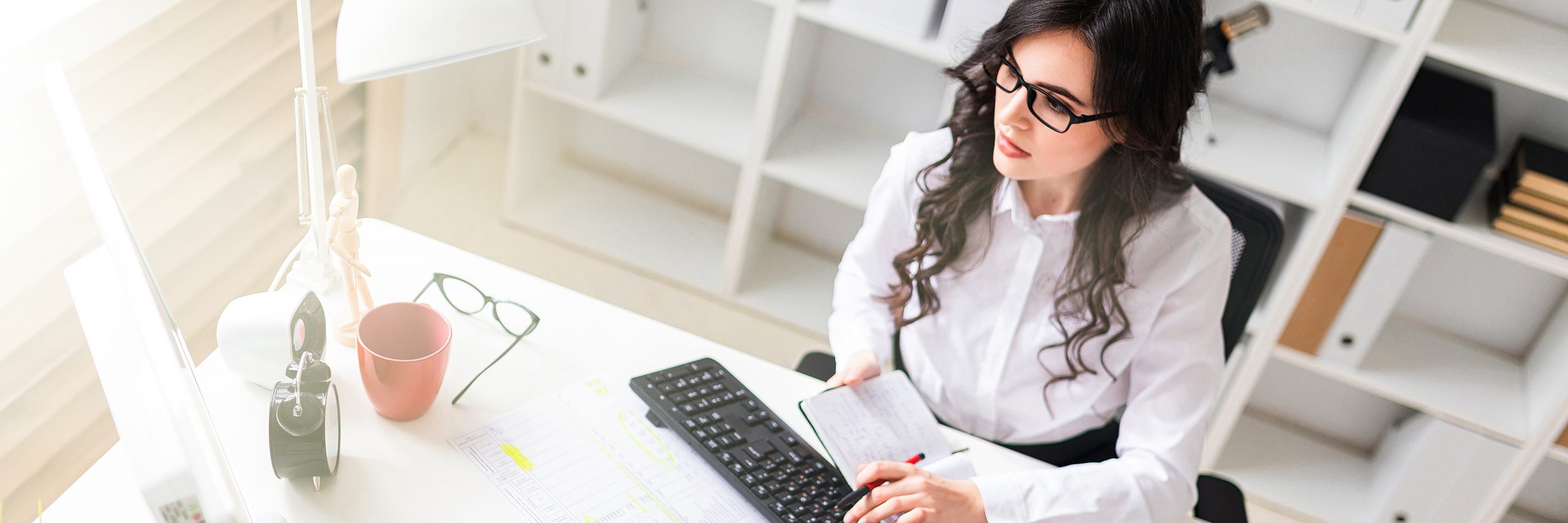 A young girl is working at the computer in the office and holds a pen and notebook in her hand.